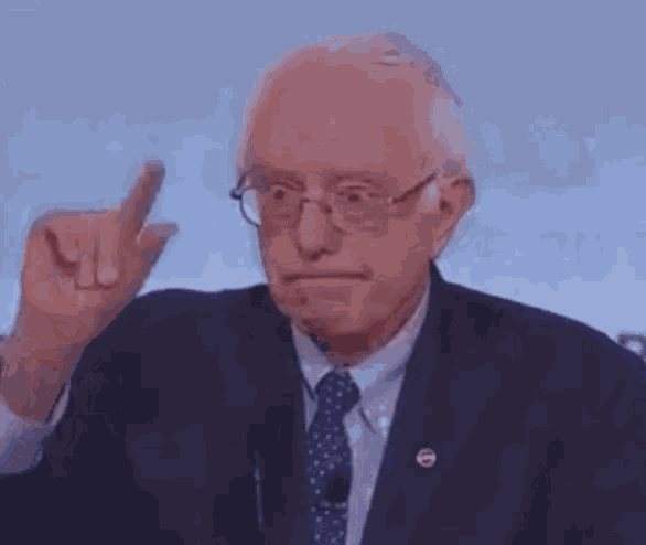 sanders is wearing glasses and a suit and tie and pointing his finger at the camera .