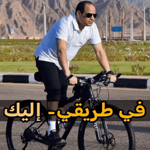a man wearing sunglasses is riding a bike on a road with arabic writing behind him