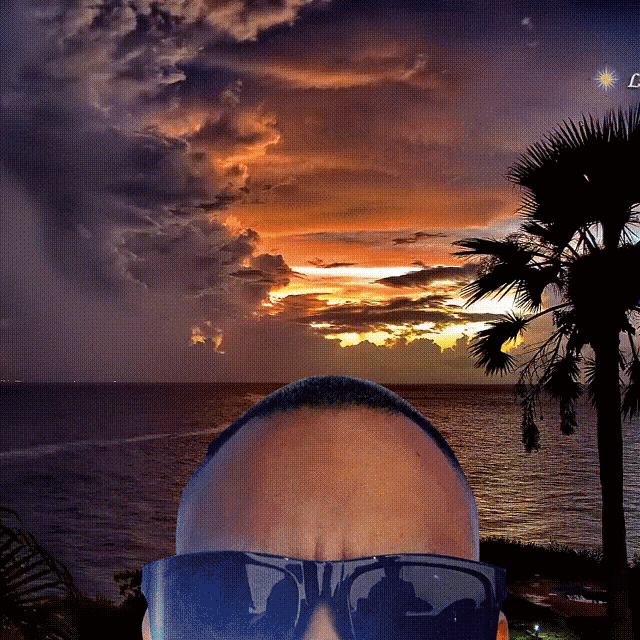 a man wearing sunglasses takes a selfie in front of a sunset over the ocean