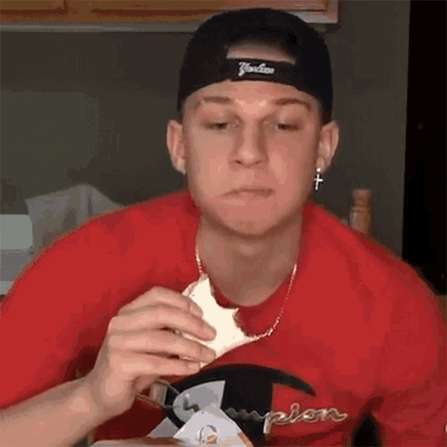 a man wearing a yankees hat and a red champion shirt is eating a sandwich