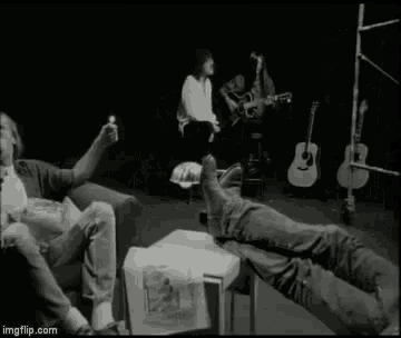 a black and white photo of a group of people sitting on a couch with their feet up in front of guitars .