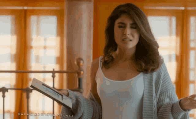 a woman in a white tank top and grey cardigan holds a book in front of a window