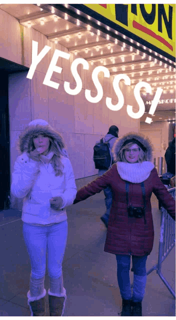 two women standing in front of a sign that says yessss