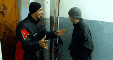 a man in a black jacket with the letter a on it talks to another man in a grey hat