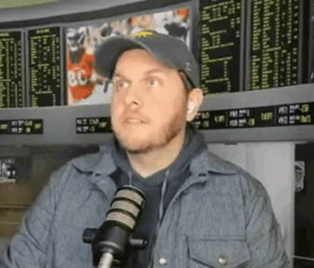 a man wearing a hat and a jacket is talking into a microphone in front of a betting board .