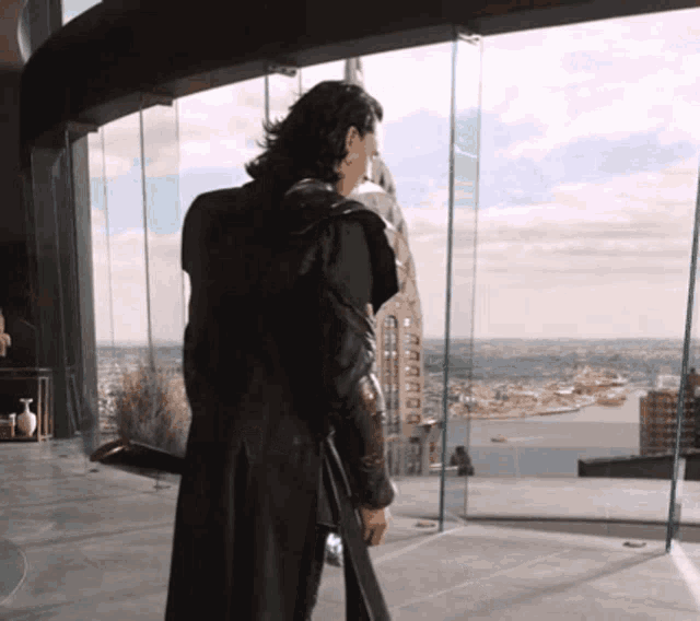 a man in a black coat stands in front of a window with a view of a city