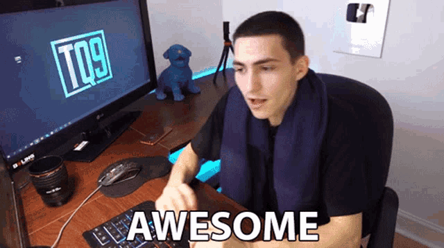 a man with a scarf around his neck is sitting at a desk in front of a computer screen that says tq9