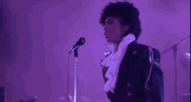prince is singing into a microphone on a stage in a purple light .