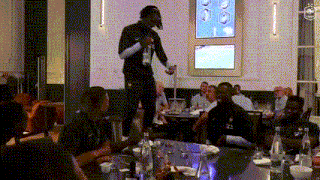 a man is standing on a table holding a microphone in front of a group of people sitting at a table .
