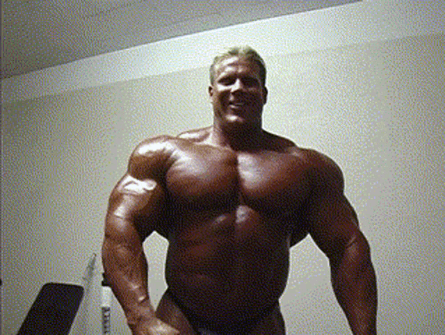 a very muscular man stands in front of a white wall and a bench