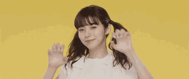 a young woman in a white shirt is making a cat face with her hands on a yellow background .