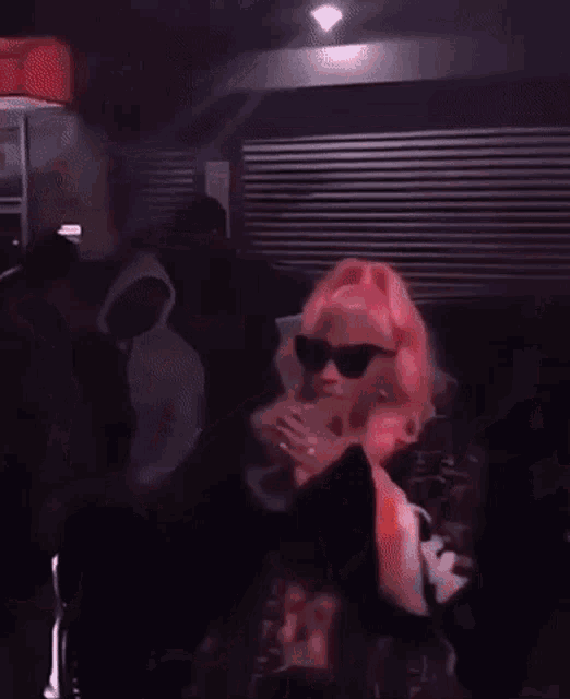 a woman with pink hair is wearing sunglasses and headphones while dancing in a dark room .
