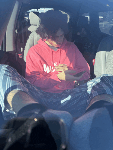 a man in a pink adidas hoodie sits in the back seat of a car looking at his phone