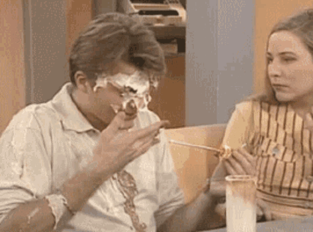 a man and a woman are sitting at a table eating food . the man has a lot of whipped cream on his face .