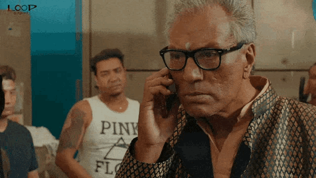 a man wearing glasses is talking on a cell phone with a man wearing a pink shirt behind him