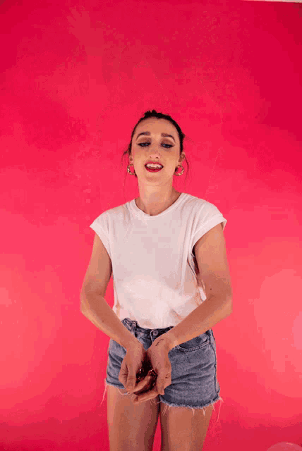 a woman in a white t-shirt and denim shorts stands in front of a pink background