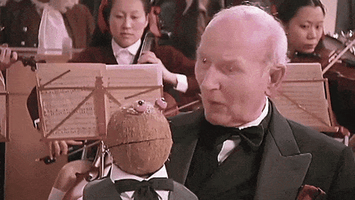 a man in a tuxedo holds a puppet in front of an orchestra playing violins