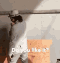 a man in a white hat is standing in front of a wall with the words " do you like it " written on it .