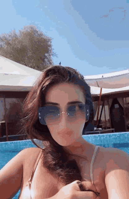 a woman in a bikini and sunglasses is taking a selfie in front of a pool .