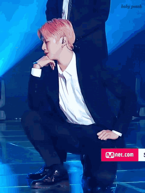 a man in a suit and white shirt is squatting down on a stage with a mnet.com sign in the foreground