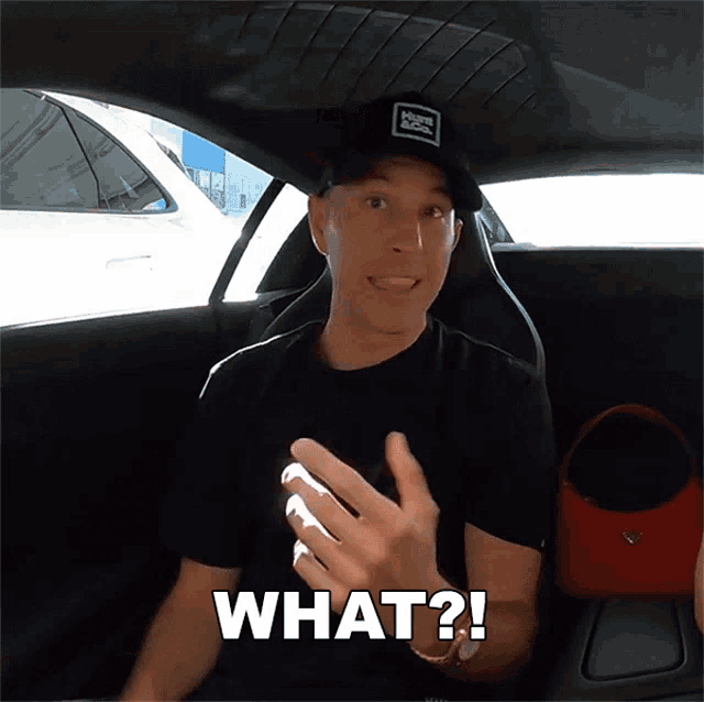 a man in a black shirt is sitting in a car and the word what is above him