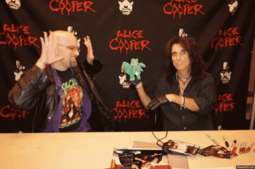 two men sitting at a table in front of a alice cooper banner