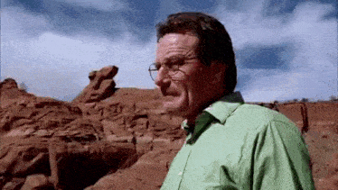 a man wearing glasses and a green shirt is standing in front of a rock formation in the desert .