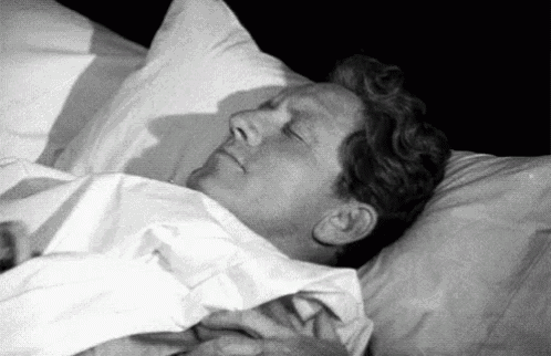 a man is sleeping in a bed with his eyes closed and his hands on his chest .