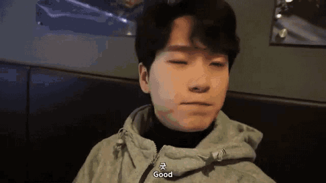 a young man wearing a hoodie is sitting at a table with his eyes closed and says `` good '' .