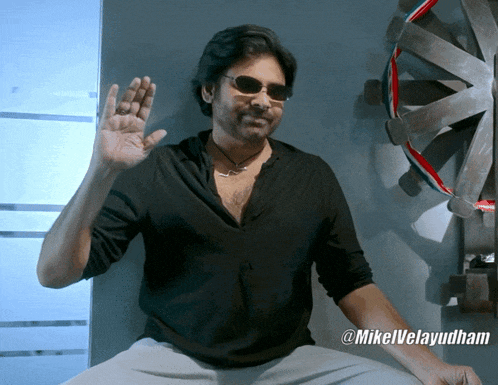 a man wearing sunglasses and a black shirt is sitting in front of a dart board and says @mikelvelayudham
