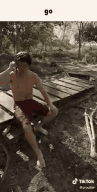 a shirtless man is sitting on a wooden bench in the woods talking on a cell phone .