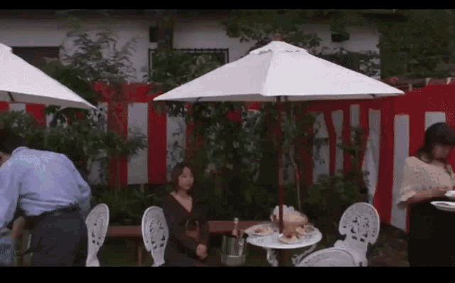 a woman sits at a table under an umbrella while a man stands behind her