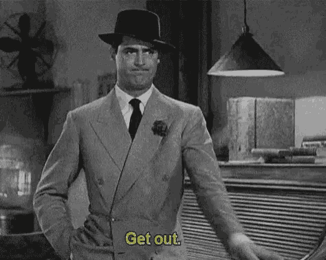 a man in a suit and hat is standing in front of a counter and says `` get out '' .