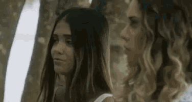 two women are standing next to each other in a forest looking at something .
