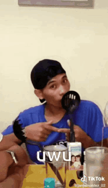 a man in a blue shirt is sitting at a table with a spoon in his mouth and the word uwu on the screen .