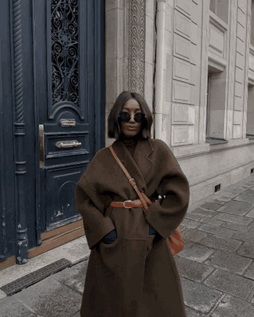 a woman wearing a brown coat and sunglasses stands in front of a building