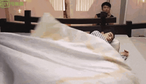 a man is laying on a bed with a white blanket while another man looks on .