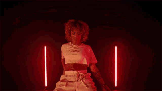 a woman in a white crop top and white shorts is dancing in front of red lights in a dark room .