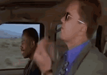 a man in a suit and sunglasses is driving a car with another man in the back seat .