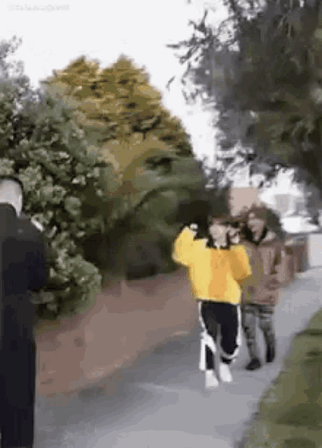 a woman in a yellow sweater is walking down a sidewalk while a man takes a picture of her .