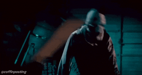 a man in a hooded jacket is standing in a dark room with a barrel in the background .