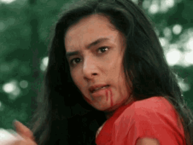 a woman with blood on her face and a red shirt is looking at the camera .