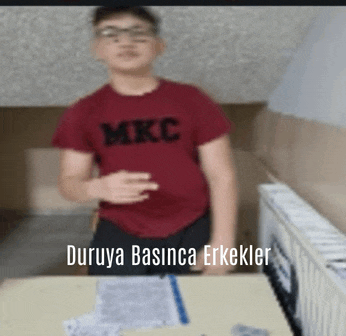 a boy wearing a red mkc shirt stands in front of a desk