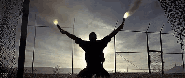a man is kneeling down with his arms outstretched and holding two flares in front of a fence .