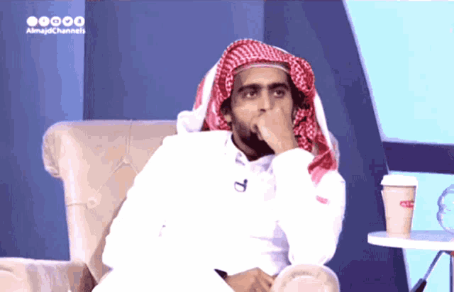 a man wearing a head scarf is sitting in a chair with a cup of coffee on a table in front of him