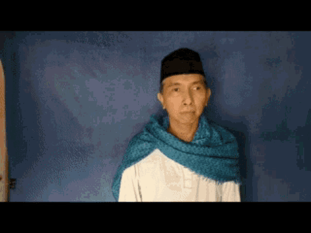 a man wearing a black hat and a blue scarf stands in front of a blue background