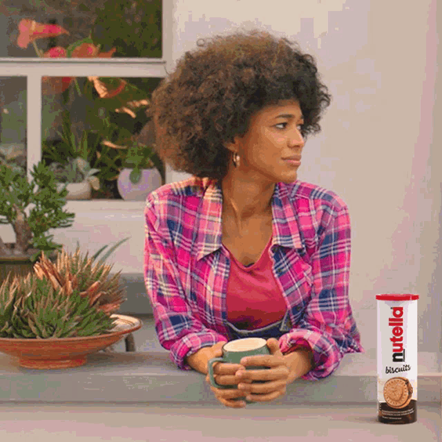 a woman in a plaid shirt holds a cup of coffee next to a can of nutella biscuits