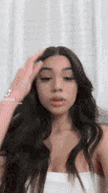 a woman with long dark hair is holding her hand to her forehead and looking at the camera .