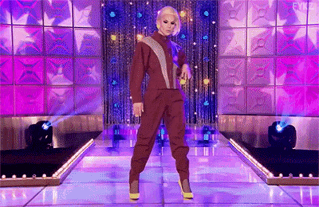 a drag queen is dancing on a stage while wearing a jumpsuit and yellow shoes .