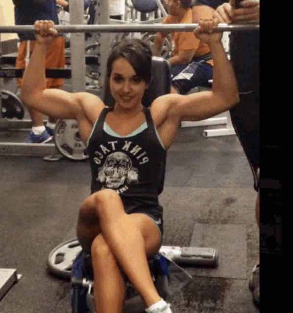 a woman wearing a black tank top that says jat miami is lifting a barbell in a gym